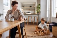 A calm parent following child product recall steps on a phone at the kitchen table while a young child plays safely nearby