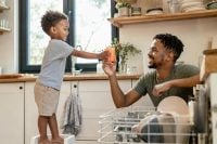A cheerful young child in a bright kitchen helping a parent put away clean dishes, illustrating age appropriate chores for kids