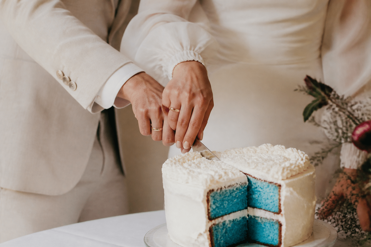 Gender reveal outfit ideas for a cake reveal — a couple's hands cutting into a cake with blue filling, both wearing neutral cream-colored outfits, soft indoor lighting