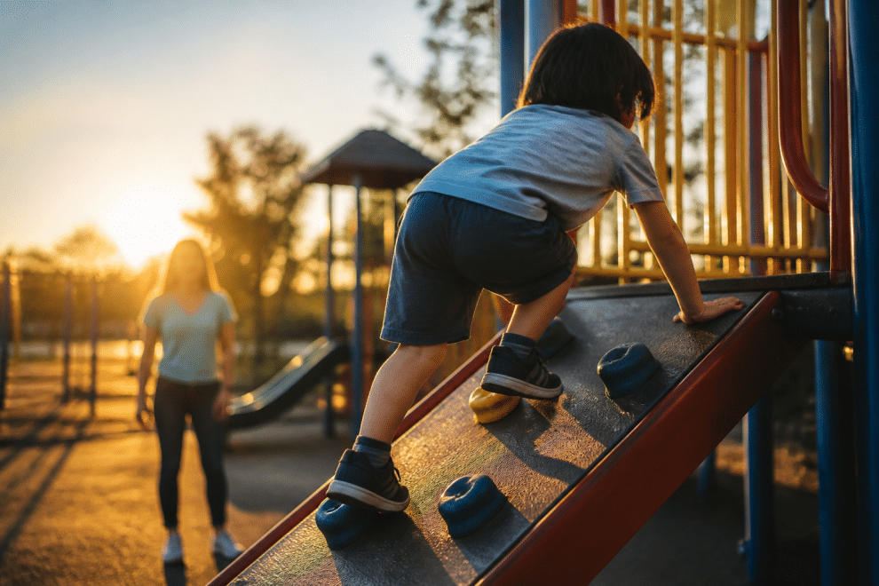 how to raise a confident child — child climbing playground equipment with parent encouraging nearby
