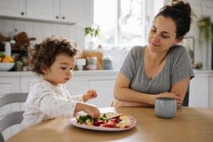 Picky eater toddler tips — a toddler at a family kitchen table exploring colorful foods on a plate, a parent nearby with a relaxed expression, warm natural light.